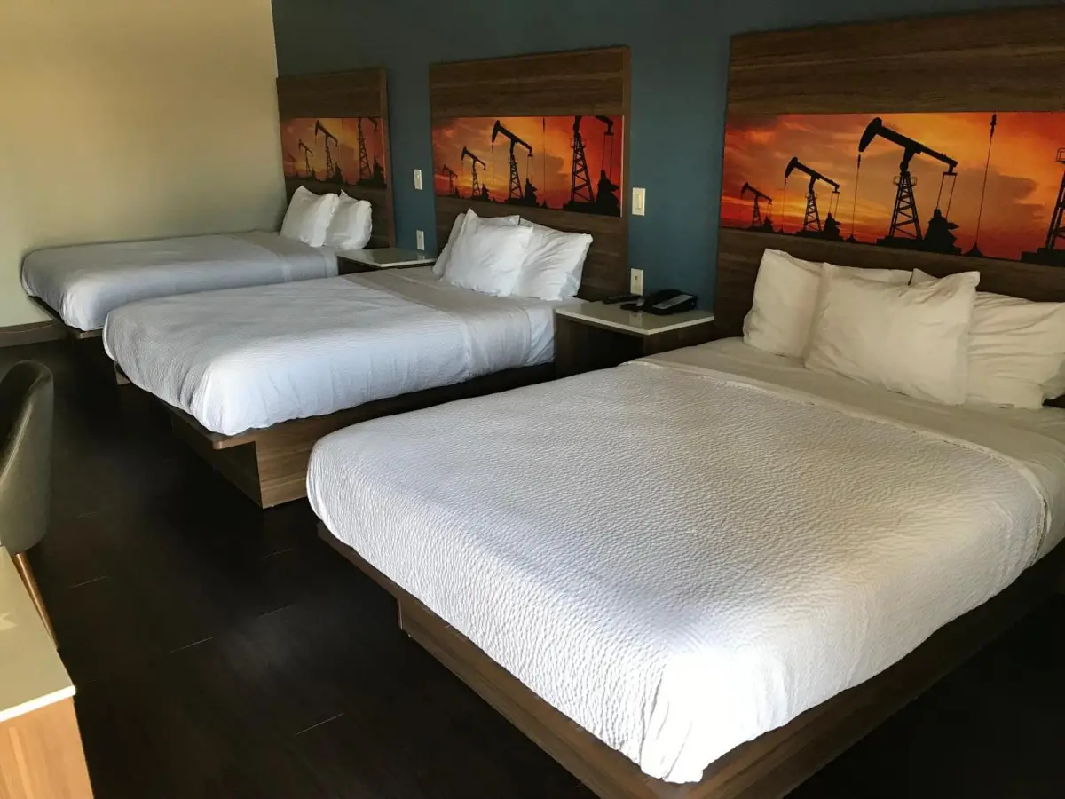 A hotel room with three beds, white bedding, and oil rig artwork on the headboards.