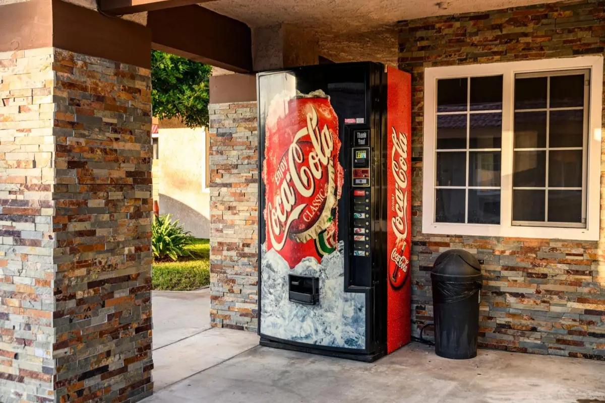 A vending machine and trash can outside a building with a stone wall and window.