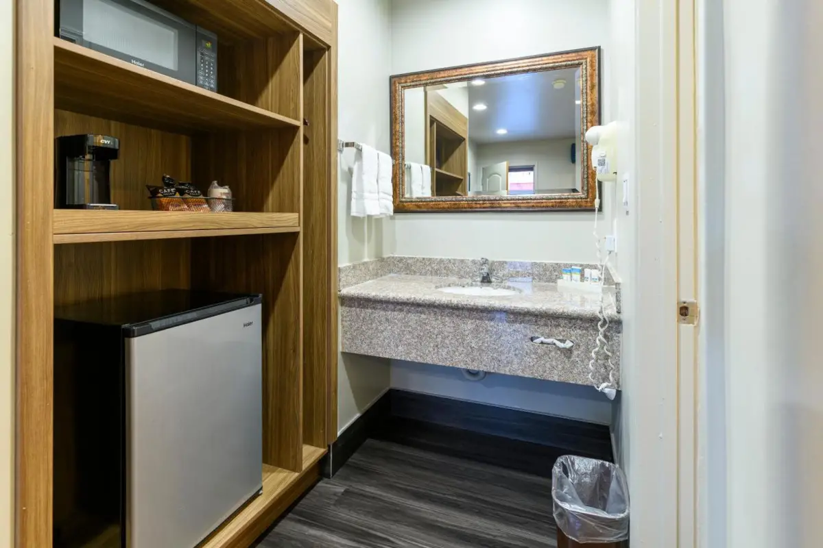 A bathroom with a granite countertop, wooden shelves, a small fridge, and a mirror
